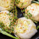 Close up of raw garlic heads with fresh spices in cast iron skillet for preparing roasted garlic recipe.