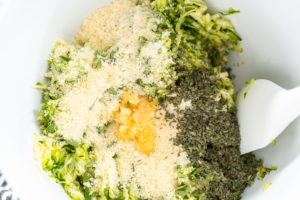 Mixing bowl with shredded zucchini, herbs, breadcrumbs, and cheese before blending.