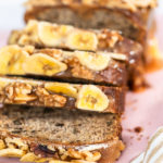 Sliced loaf of banana nut bread drizzled homemade caramel on a pink cutting board.