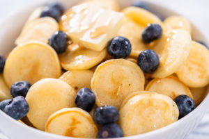 Mini pancake cereal - Arina Photography Cooked mini pancakes topped with fresh blueberries in a white bowl – final serving of mini pancake cereal with blueberries and syrup for a sweet breakfast.