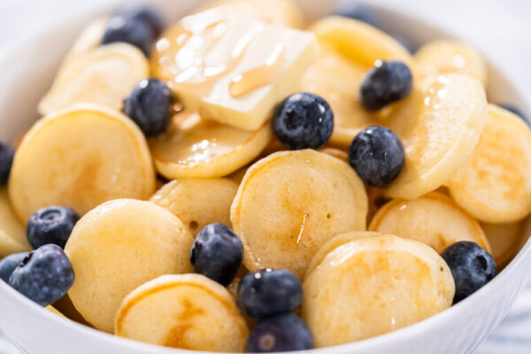 Mini pancake cereal - Arina Photography Cooked mini pancakes topped with fresh blueberries in a white bowl – final serving of mini pancake cereal with blueberries and syrup for a sweet breakfast.