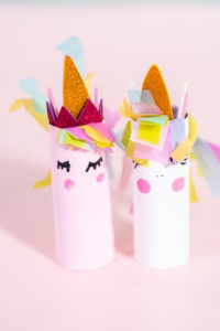 Two colorful unicorn crafts made from toilet paper rolls, painted pink and white with hand-drawn eyes, blush, and gold glitter horns, decorated with pastel tissue paper manes, displayed on a light pink background.