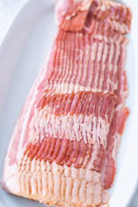 Cooking bacon strips - Arina Photography A close-up stack of raw bacon strips on a white plate.