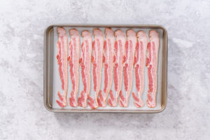 Cooking bacon strips - Arina Photography Uncooked bacon strips neatly arranged on a parchment-lined baking sheet.