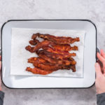 Hands holding a tray of crispy, baked bacon strips on parchment paper.