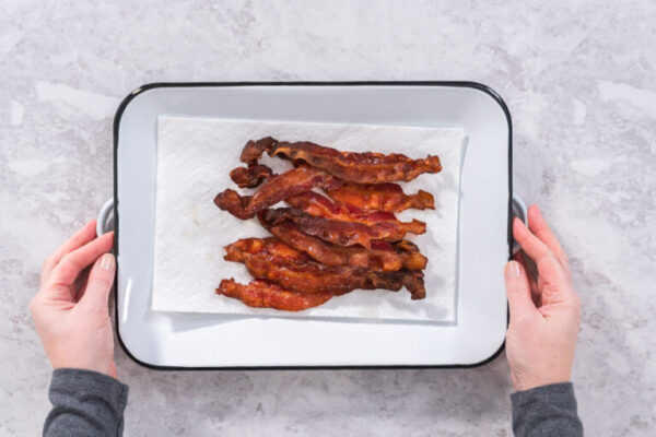 Hands holding a tray of crispy, baked bacon strips on parchment paper.