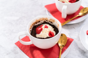 Chocolate mug cake - Arina Photography Overhead shot of single Nutella mug cake decorated with whipped cream and red candy hearts, placed on a red napkin with gold spoon.