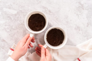 Chocolate mug cake - Arina Photography Hands holding two white mugs filled with freshly microwaved chocolate mug cakes on a marble counter.