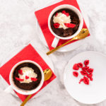 Top-down view of two finished Nutella mug cakes topped with whipped cream and red candy hearts, served on red napkins.