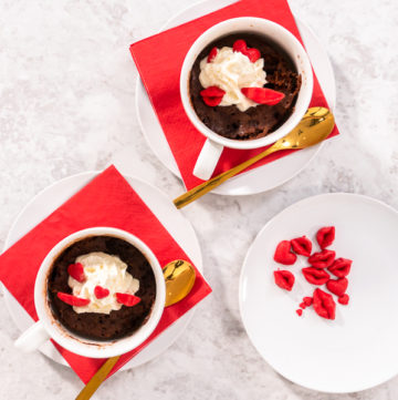 Chocolate mug cake - Arina Photography Top-down view of two finished Nutella mug cakes topped with whipped cream and red candy hearts, served on red napkins.