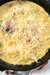 Spinach and ham frittata - Arina Photography Shredded cheese and cubed ham layered over egg mixture in a skillet before baking.