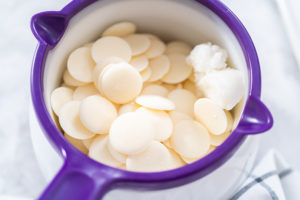 Close-up of white candy melts and a scoop of coconut oil inside a purple candy melt pot, ready to be melted for chocolate-covered pretzels.