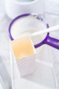 Tall paper container filled with melted white candy coating, ready for dipping pretzel rods, with a candy melt pot and spatula in the background.