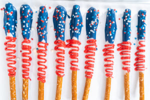 Red, white, and blue chocolate-covered pretzel rods decorated with patriotic star sprinkles and red candy drizzle, arranged on parchment paper.