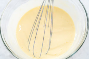 Whisk blending a smooth, pale yellow egg mixture in a glass bowl during Bundt cake preparation.