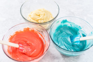 Three bowls of red, white, and blue cake batter with spatulas, ready to layer into a patriotic Bundt cake.