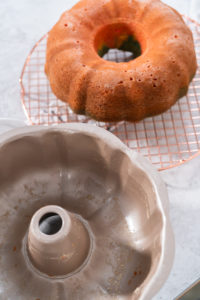 Freshly baked Bundt cake cooling on a wire rack beside an empty, greased Bundt pan.