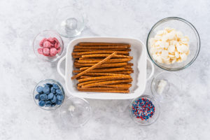Overhead view of ingredients for patriotic chocolate-covered pretzel rods, including pretzel sticks, white candy melts, red and blue melts, coconut oil, and red-white-blue sprinkles in glass bowls on a marble countertop.