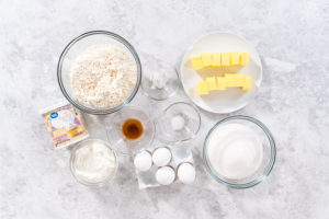 Overhead view of Bundt cake ingredients including flour, butter, eggs, sugar, sour cream, vanilla, salt, baking powder, and frosting on a marble surface.