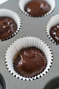Close-up of chocolate cupcake batter layered over graham cracker crust in a white paper cupcake liner, ready to be baked.