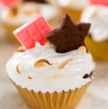 Toasted s’mores cupcake topped with swirled marshmallow meringue frosting, pink chocolate bar, and star-shaped chocolate graham cracker in a gold foil liner.