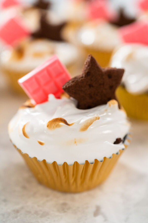 S'mores cupcakes - Arina Photography Toasted s’mores cupcake topped with swirled marshmallow meringue frosting, pink chocolate bar, and star-shaped chocolate graham cracker in a gold foil liner.
