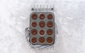 Freshly baked chocolate cupcakes in a muffin tin lined with white and gold foil cupcake liners, cooling on a metal rack with a striped kitchen towel.