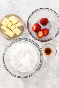 Ingredient prep: bowls of butter, sliced strawberries, powdered sugar, and vanilla extract on a counter.