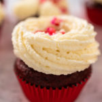 Freshly baked velvet cupcakes with white chocolate ganache frosting decorated with sprinkles.