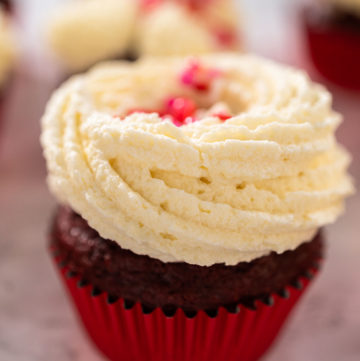 Freshly baked velvet cupcakes with white chocolate ganache frosting decorated with sprinkles.