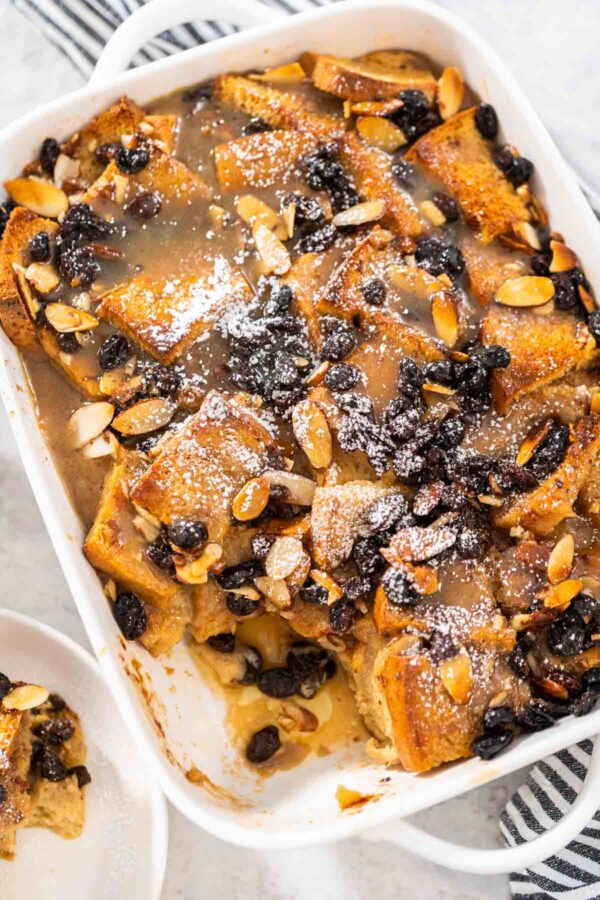 Serving freshly baked bread pudding with raising and almond slivers in a white casserole dish.