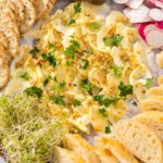 Butter board with vegetables and bread on a round wood cutting board.