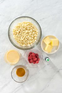 Overhead view of measured ingredients in glass bowls on a marble surface, including white chocolate chips, cubes of butter, sweetened condensed milk, vanilla extract, red candy melts, and a small bottle of green food coloring, prepared for making festive Grinch Fudge.