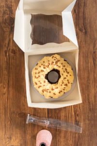 Flat lay. Carefully placing the gingerbread bundt cake, adorned with salted caramel frosting and gingerbread sprinkles, into a white paper bundt cake box with a clear window for gifting.