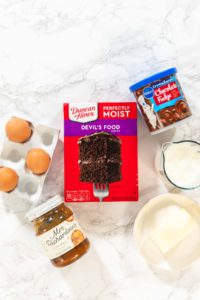Baking Chocolate Cupcakes with Decadent Chocolate Frosting - Arina Photography Top-down view of ingredients for fancy chocolate cupcakes: box of Duncan Hines Devil’s Food cake mix, Pillsbury chocolate fudge frosting, Mrs. Richardson’s caramel sauce, four eggs in a ceramic holder, a glass measuring cup with milk, and a bowl with two sticks of butter—arranged on a white marble background.