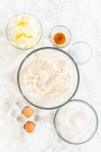 Overhead view of neatly arranged ingredients in glass bowls for sugar cookie dough, including flour, butter, sugar, egg, and vanilla.