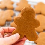 Close-up of a hand holding a baked gingerbread man cookie over a cooling rack filled with cookies.