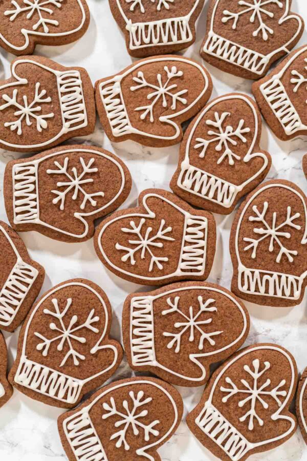 Beautifully Decorated Mitten Cookies with Royal Icing Designs - Arina Photography Overhead view of multiple mitten-shaped chocolate cookies decorated with white royal icing snowflakes and ribbed cuffs, arranged on a marble background.
