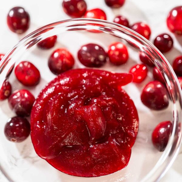 835-05010 - Arina Photography Glass bowl filled with smooth cranberry puree, surrounded by fresh whole cranberries on a white surface.