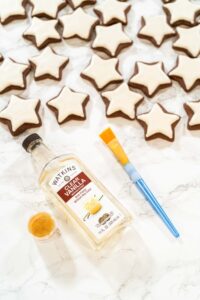 Perfectly Icing Star Cookies with Royal Icing - Arina Photography Flat lay of iced chocolate star cookies with decorating tools including Watkins clear vanilla extract, edible gold dust, and a paintbrush—perfect setup for adding shimmer to holiday cookies.
