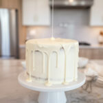 Smooth white chocolate ganache being poured over a frosted layer cake on a white cake stand in a bright kitchen setting.