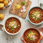 071-featured image23 - Arina Photography Bowls of hearty turkey chili topped with shredded cheese, radish slices, green onions, and cilantro, served with golden cornbread muffins on a rustic white table.